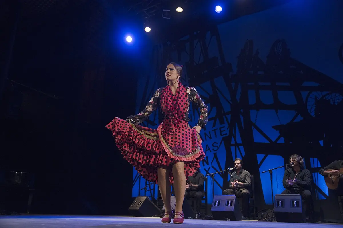 Una bailaora flamenca con un vestido rojo de lunares permanece de pie con una mano sobre la falda en un escenario iluminado en azul, con cantaores y guitarristas sentados detrás de ella