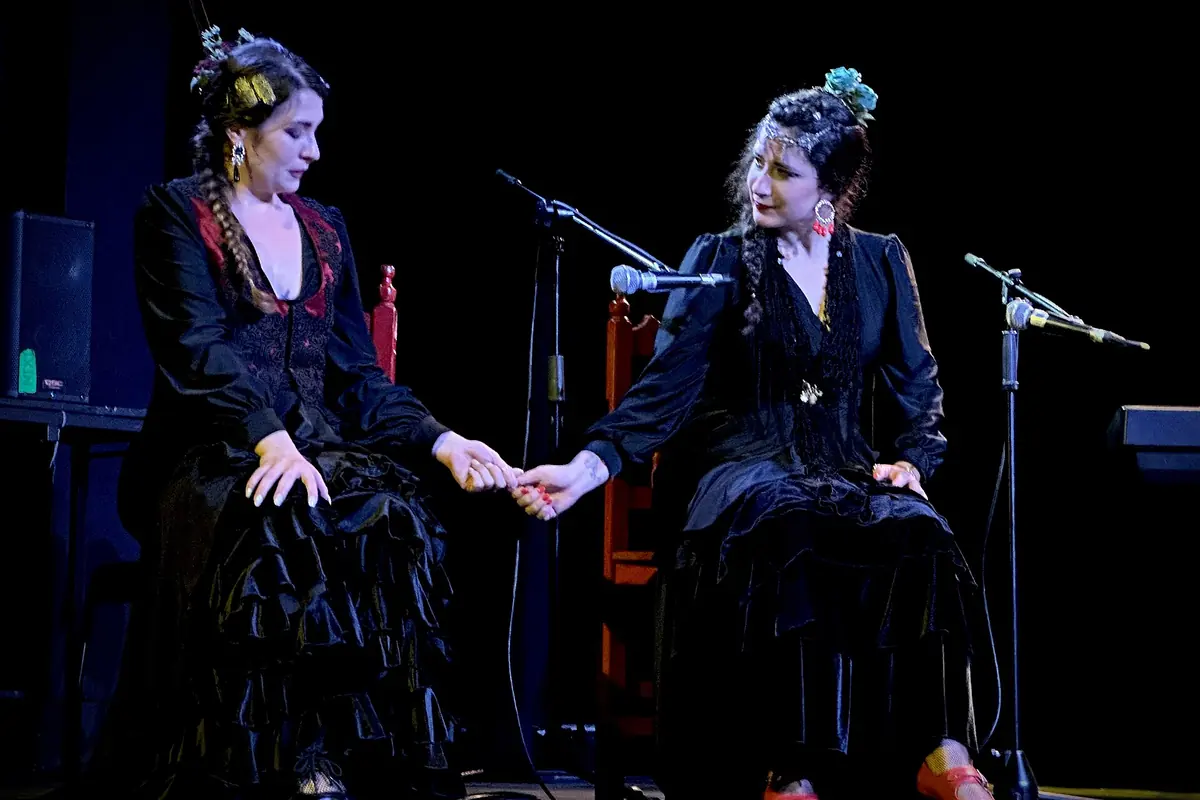 Dos mujeres con vestidos negros y flores en el pelo permanecen sentadas de la mano sobre un escenario oscuro junto a unos micrófonos