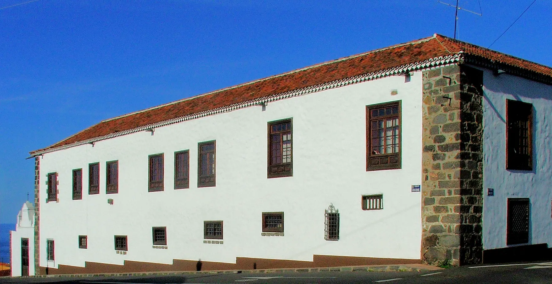 La larga fachada encalada de la Casa Ábaco bajo un cielo azul, con tejado de teja roja, esquinas de piedra oscura y ventanas de madera marrón