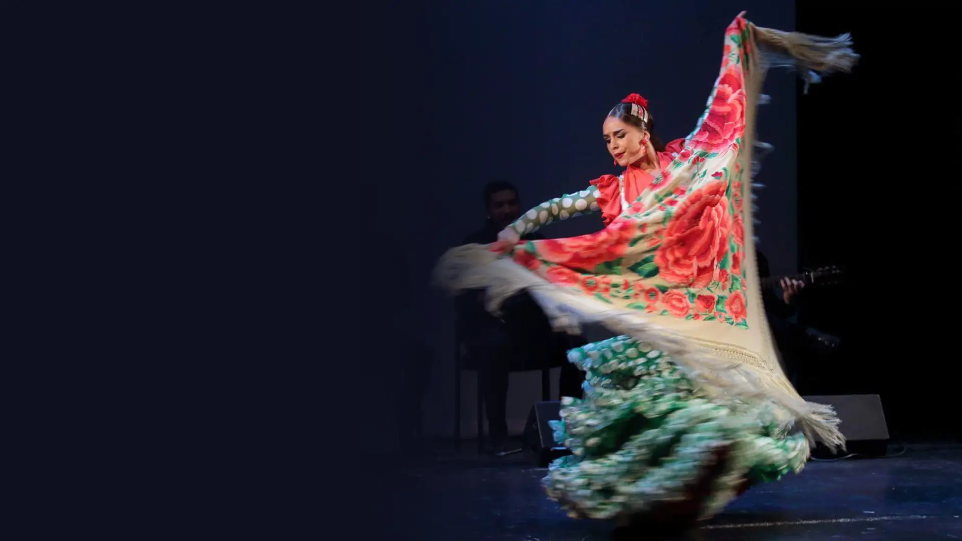 Una bailaora flamenca con un vestido de volantes de lunares verdes hace ondear un gran mantón crema bordado con rosas rojas sobre un escenario oscuro, con los músicos sentados detrás de ella