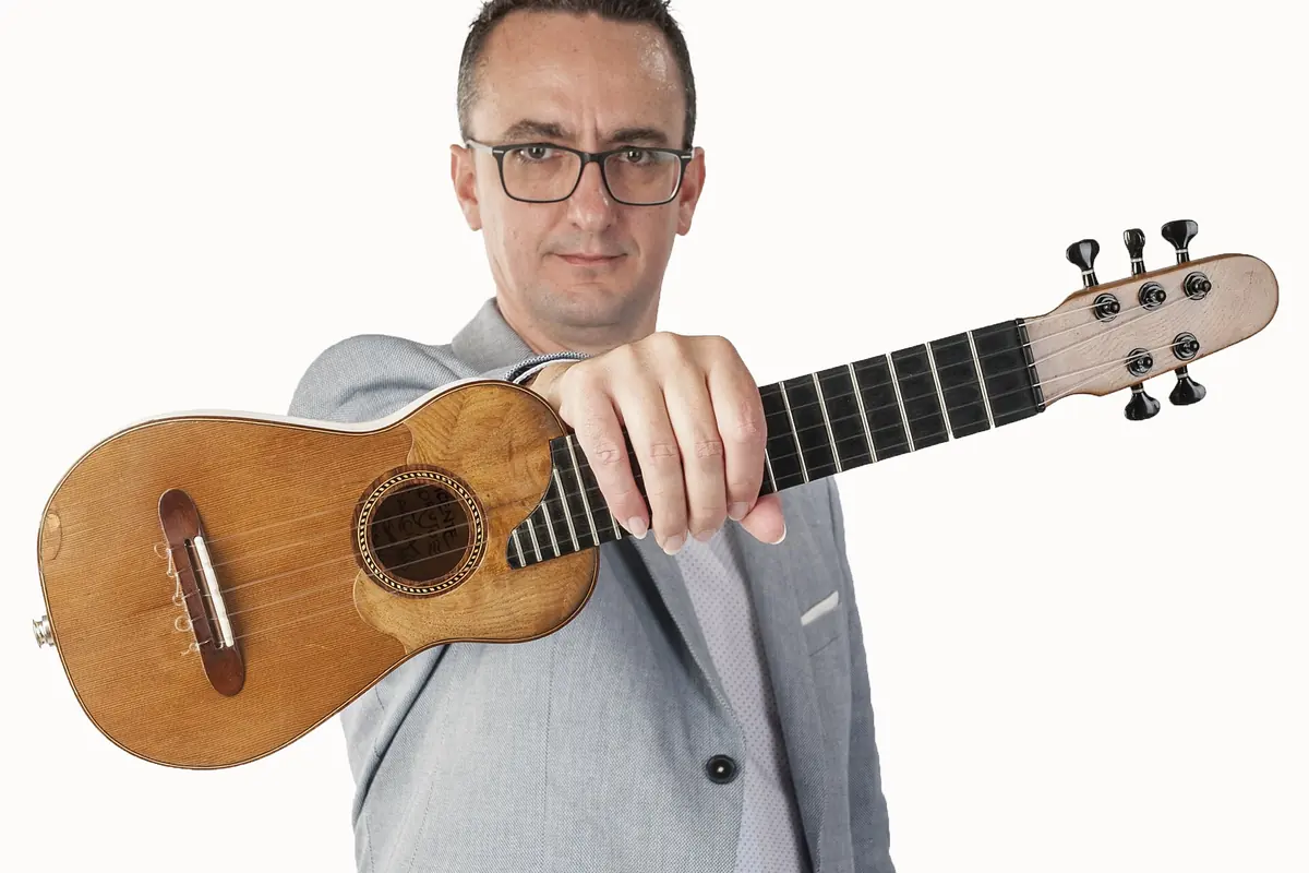 Mężczyzna w okularach i szarej marynarce trzyma przed kamerą timple, mały pięciostrunowy instrument kanaryjski z jasną główką