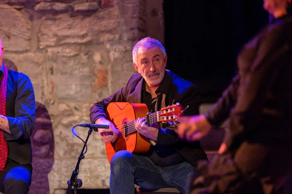 Un guitarrista flamenco de pelo canoso toca una guitarra dorada frente a un micrófono mientras el brazo de una bailaora se mueve en primer plano